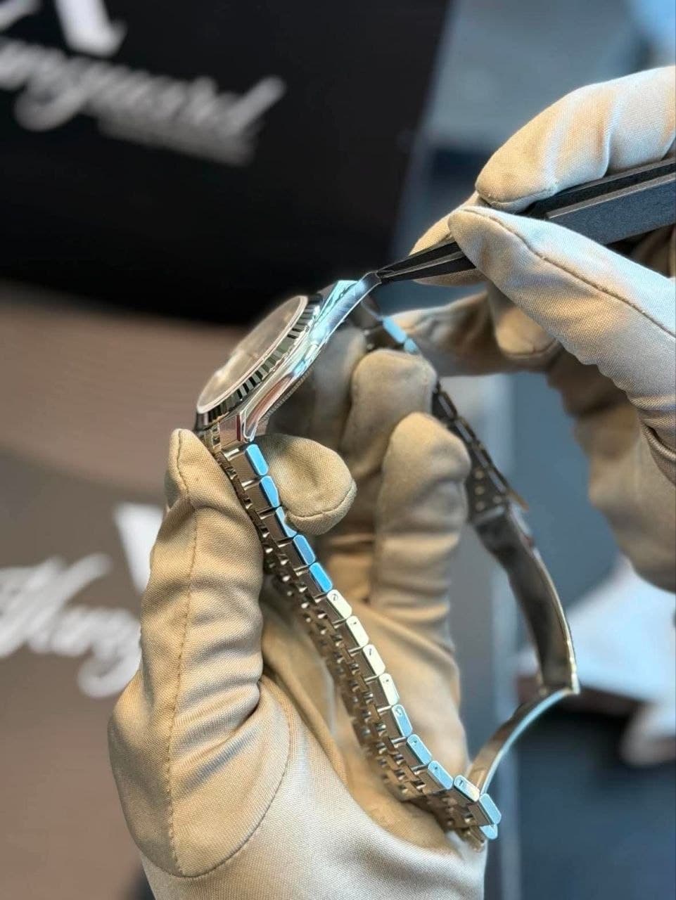 A steel Rolex Datejust held side-on in gloved hands while tweezers lay protective film along the edge of its Jubilee bracelet