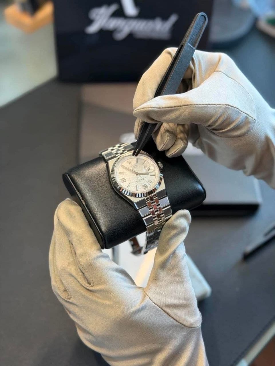 Gloved hands lifting the edge of a protective film with tweezers from the case of a steel Rolex Datejust, the watch resting on a black cushion