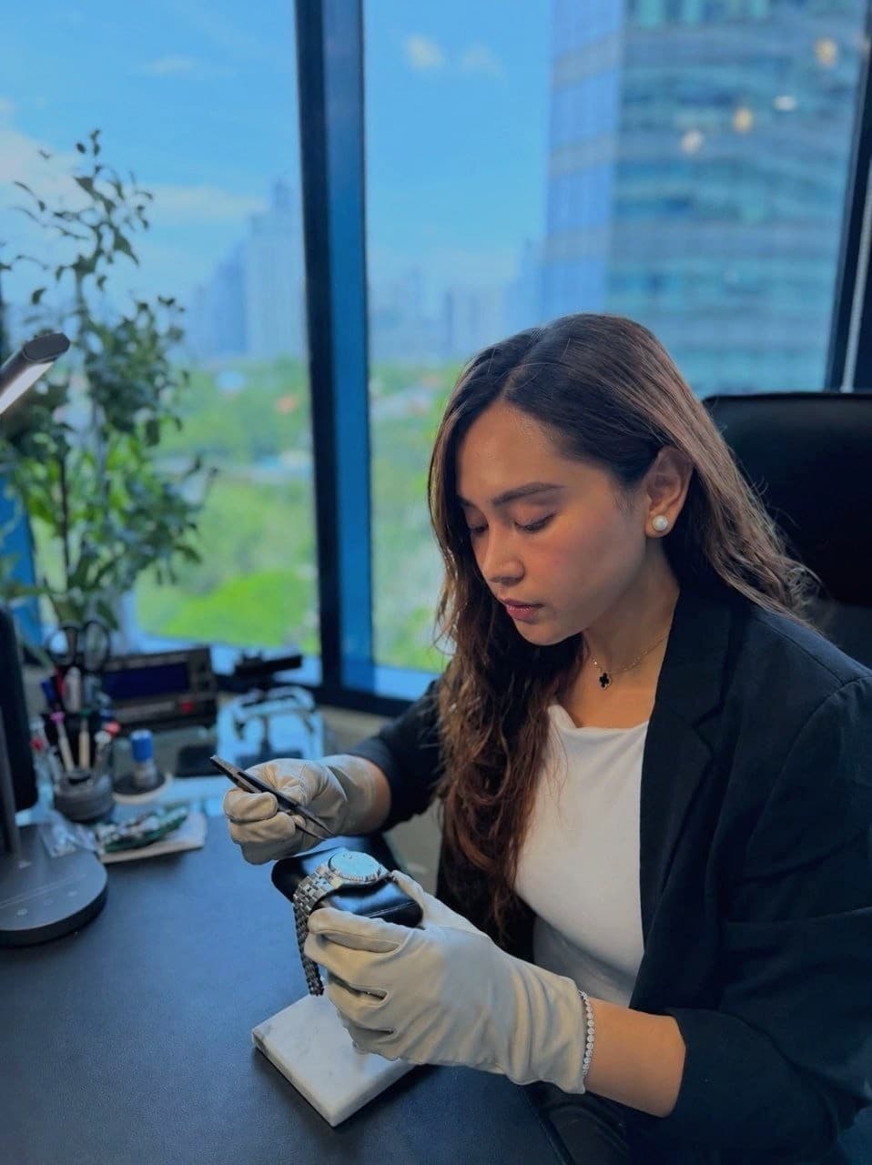 Gloved hands at the bench, tweezers in one hand, working on a steel Rolex Datejust held upright on a stand, the city skyline through the window behind