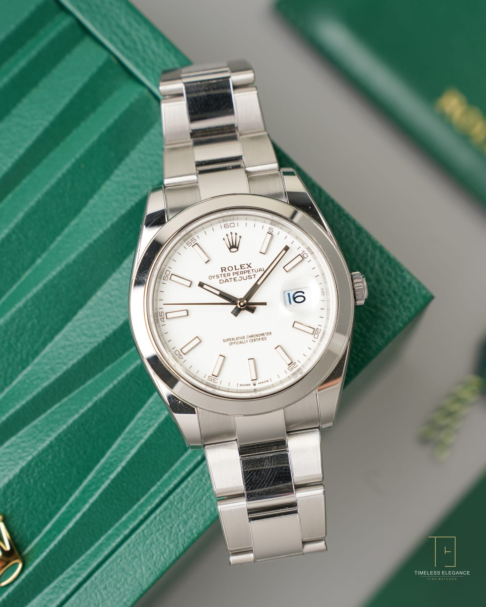A steel Rolex Datejust with white dial on an Oyster bracelet, beside its green Rolex case